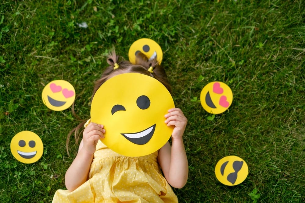 A Child Hides His Face Behind A Yellow Winking Emoticon